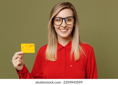 Young smiling happy rich fun cool woman she wears red shirt casual clothes glasses hold in hand mock up of credit bank card isolated on plain pastel green background studio portrait. Lifestyle concept