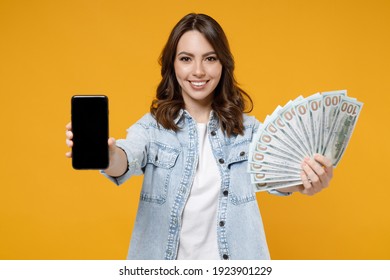 Young smiling happy rich woman in denim shirt white t-shirt showing mobile cell phone, blank screen workspace area fan of cash money in dollar banknotes isolated on yellow background studio portrait.