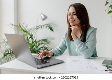 Young Smiling Happy Satisfied Employee Business Woman In Casual Blue Shirt Hold Pen Sit Work At Workplace White Desk With Laptop Pc Computer At Light Modern Office Indoors. Achievement Career Concept