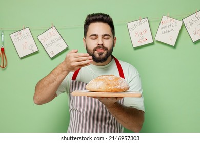 Young Smiling Happy Satisfied Male Chef Confectioner Baker Man 20s In Striped Apron Hold Warm Homemade Bread Sniff Scent Isolated On Plain Pastel Light Green Background Studio. Cooking Food Concept