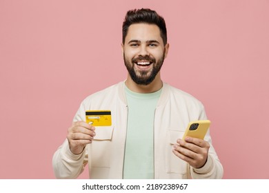 Young Smiling Happy Satisfied Man In Trendy Jacket Shirt Using Mobile Cell Phone Hold Credit Bank Card Doing Online Shopping Order Delivery Booking Tour Isolated On Plain Pastel Light Pink Background