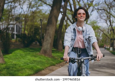 Young smiling happy sporty woman 20s wearing jeans clothes headphones riding bicycle bike on sidewalk in city spring park outdoors, look aside. People active urban healthy lifestyle cycling concept - Shutterstock ID 1997465816