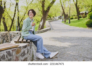 Young Smiling Happy Student Woman 20s In Casual Green Jacket Jeans Sitting On Bench In City Spring Park Outdoors Resting Eat Apple Fruit Look Aside. People Vegeterian Healthy Urban Lifestyle Concept.