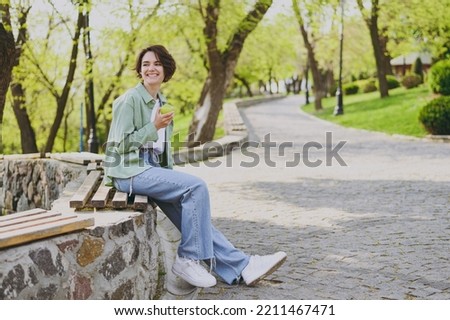 Young smiling happy student woman 20s in casual green jacket jeans sitting on bench in city spring park outdoors resting eat apple fruit look aside. People vegeterian healthy urban lifestyle concept.