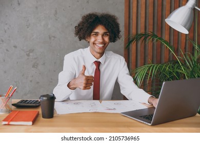 Young smiling happy successful african american employee business man 20s in classic shirt tie sit work at white office desk with pc laptop computer show thumb up inside Achievement career concept.