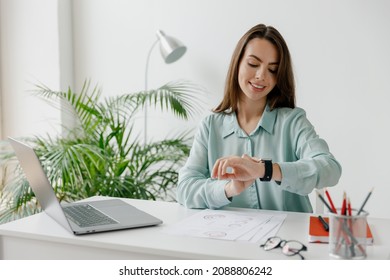 Young smiling happy successful employee business woman 20s in blue shirt look at smartwatch time sit work at workplace white desk with laptop pc computer at office indoors. Achievement career concept - Shutterstock ID 2088806242