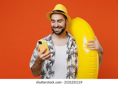 Young Smiling Happy Tourist Man In Beach Shirt Hat Hold Inflatable Ring Hold In Hand Use Mobile Cell Phone Isolated On Plain Orange Background Studio Portrait. Summer Vacation Sea Rest Sun Tan Concept