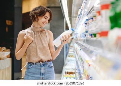 Young smiling happy woman 20s in casual clothes backpack shopping at supermaket grocery store buy choose dairy produce take milk read shelf life inside hypermarket Purchasing gastronomy food concept.