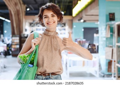 Young Smiling Happy Woman 20s In Casual Clothes Shopping At Supermaket Store With Green Eco Bag Show Thumb Up Gesture Produce Inside Hypermarket. People Lifestyle Purchasing Gastronomy Food Concept