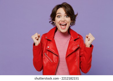 Young smiling happy woman 20s wear red leather jacket doing winner gesture celebrate clenching fists say yes isolated on plain pastel light purple background studio portrait. People lifestyle concept