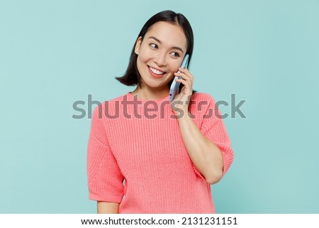 Young smiling happy woman of Asian ethnicity 20s in pink sweater talk speak on mobile cell phone conducting pleasant conversation isolated on pastel plain light blue color background studio portrait.