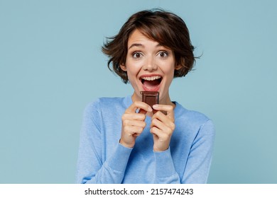 Young smiling happy woman in casual sweater hold sweet pink cream donuts biting chocolate bar look camera isolated on plain pastel light blue background studio portrait. People lifestyle food concept
