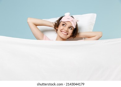 Young smiling happy woman in pajamas jam sleep eye mask rest relax at home lie in bed on pillow under blanket hold hand behind neck isolated on pastel blue background Good mood night bedtime concept.