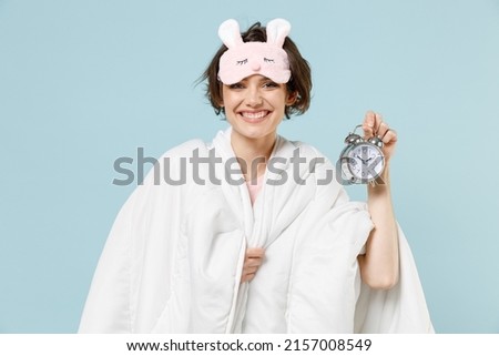 Young smiling happy woman in pajamas jam sleep eye mask rest relax at home wrap covered under blanket duvet hold clock alarm isolated on pastel blue background studio Good mood night bedtime concept