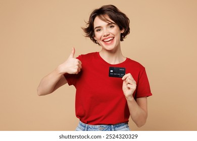 Young smiling happy woman she wear red t-shirt casual clothes hold in hand mock up of credit bank card show thumb up isolated on plain pastel light beige background studio portrait. Lifestyle concept - Powered by Shutterstock