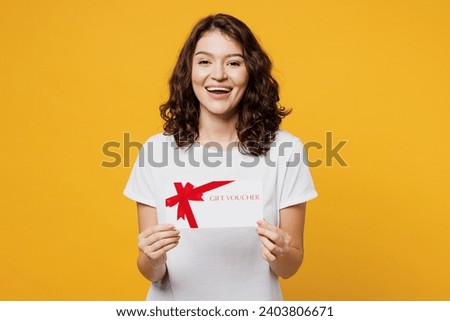 Young smiling happy woman she wear white blank t-shirt casual clothes hold gift certificate coupon voucher card for store isolated on plain yellow orange background studio portrait. Lifestyle concept