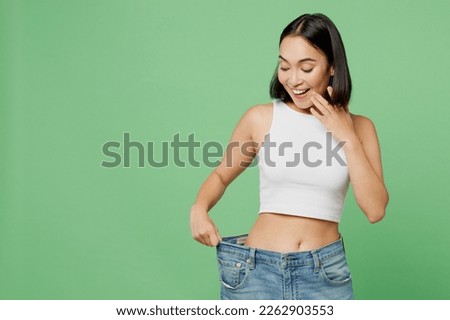 Young smiling happy woman wears white clothes show loose pants on waist after weightloss isolated on plain pastel light green background. Proper nutrition healthy fast food unhealthy choice concept