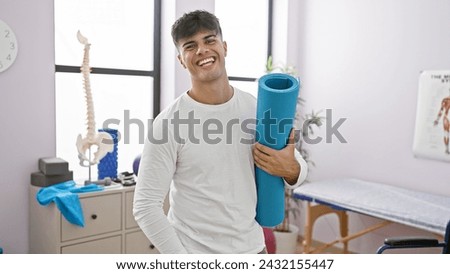 Young, smiling hispanic guy packing fitness with confidence, sporty man with yoga mat at rehab clinic, embracing athletic lifestyle for a fitter him
