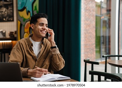 Young smiling hispanic man student sitting at the cafe table with laptop computer indoors, talking on mobile phone, noting - Powered by Shutterstock