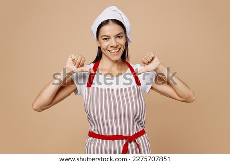 Young smiling housewife housekeeper chef cook baker latin woman wear striped apron toque hat point index finger on herself showoff isolated on plain pastel light beige background. Cooking food concept