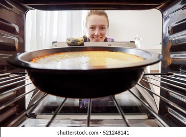 Young Smiling Housewife Looking At Cheesecake Into Oven In Kitchen. View From Inside Of The Oven. Woman Holding The Oven Door.