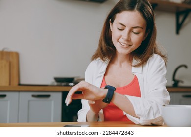 Young smiling housewife woman wear casual clothes look at smartwatch check time eat breakfast muesli cereals with milk fruit in bowl cooking food in light kitchen at home alone. Healthy diet concept - Powered by Shutterstock