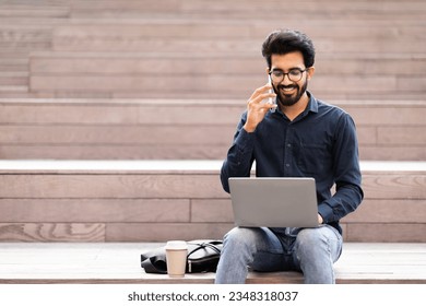 Young smiling indian man entrepreneur smiling browsing on laptop while working remotely and speaking on smartphone in street, sitting on concrete staircase, drinking takeaway coffee, copy space - Powered by Shutterstock