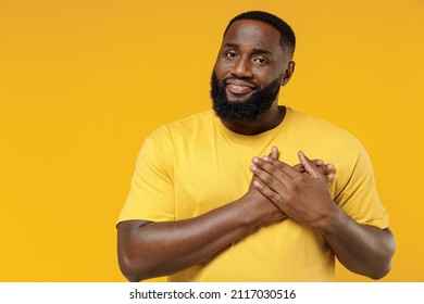 Young smiling kind-hearted happy black man 20s wearing bright casual t-shirt put folded hands on heart look camera isolated on plain yellow color background studio portrait. People lifestyle concept