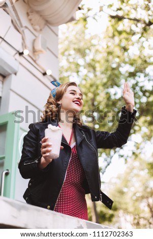 Young smiling lady in leather jacket standing with milkshake in hand and happily looking aside while waving to someone
