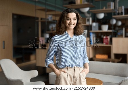Young smiling latin girl college student or teacher looking at camera standing in university campus. Happy hispanic millennial woman professional posing in modern coworking creative office space.