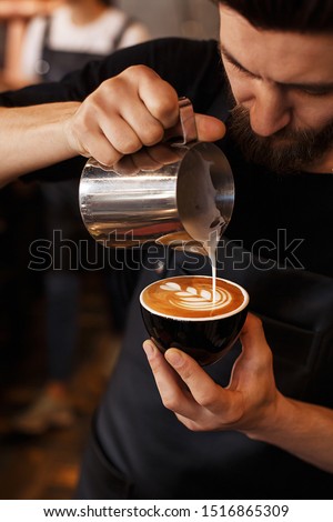 Young smiling male barista making cappuccino. Pouring milk for prepare cup of coffee. Latte art. Morning cup of coffee in café. Brewing coffee. Coffee shop concept.