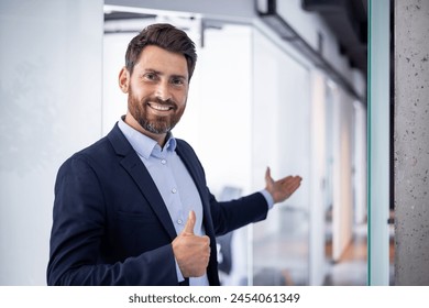 A young smiling male businessman, the founder of the company stands in a suit and invites to the office with a hand gesture of welcome and shows the super finger. - Powered by Shutterstock