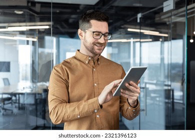 A young smiling male businessman in glasses is standing in the office and using a tablet. typing, chatting, searching for information. - Powered by Shutterstock