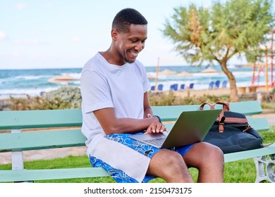 Young Smiling Male Freelancer Relaxing On Beach, Sitting On Bench Using Laptop