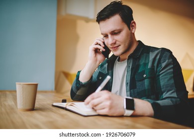 Young Smiling Male Job Seeker Having Conversation On Mobile Phone And Taking Notes In Planner Sitting At Table With Glass Of Hot Drink In Cafeteria