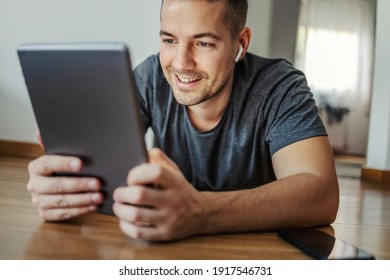A young smiling male person with wireless headphones in his ears is interested in watching a series or movie on a digital tablet. His phone was set aside on the side of the table. Relax online time - Shutterstock ID 1917546731