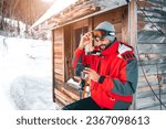 Young smiling male skier using smart phone in front of log cabin. Fashionable man in full ski equipment checking his mobile phone on vacation.