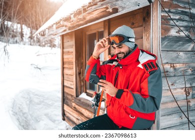 Young smiling male skier using smart phone in front of log cabin. Fashionable man in full ski equipment checking his mobile phone on vacation. - Powered by Shutterstock