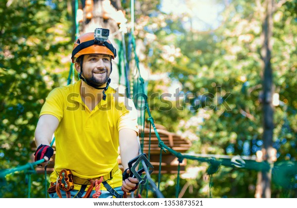 Young smiling man with action camera, in protective\
gear, climb rope trail, bridge on high trees. Rope adventure park\
with obstacles and ziplines. Extreme rest and summer activities\
concept. 