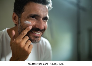 An Young Smiling Man Is Applying After Cleaning His Face Day Or Night Cream To Take Care Of Skin Behind A Mirror In A Bathroom.