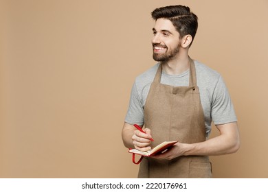 Young smiling man barista barman employee wear brown apron work in coffee shop write down order in notebook look aside isolated on plain pastel light beige background. Small business startup concept - Shutterstock ID 2220176961