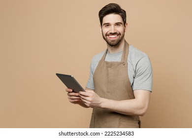 Young smiling man barista barman employee wear brown apron work in coffee shop hold use tablet pc computer look camera isolated on plain pastel light beige background. Small business startup concept - Shutterstock ID 2225234583