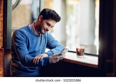 Young Smiling Man With Blue Sweater Looking At His Smartphone In A Modern Pub. Bearded Guy With Modern Hairstyle Drinking A Cola.