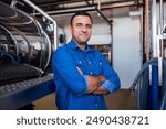 A young smiling man in a denim shirt at the cheese factory. An attractive worker near a milk and a butter making machine. An engineer for the maintenance of a cheese production line. Dairy equipment