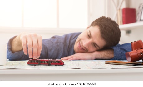 Young Smiling Man Dreaming About Travel Around World, Playing With Toy Train On Map And Choosing Route Of Vacation Trip, Copy Space