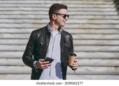 Young Smiling Man In Leather Jacket Eyeglasses Walk In City Downtown Go Down Concrete Steps Hold Mobile Cell Phone Takeaway Craft Paper Cup Drink Coffee To Go Look Aside Concept Of Urban Lifestyle.