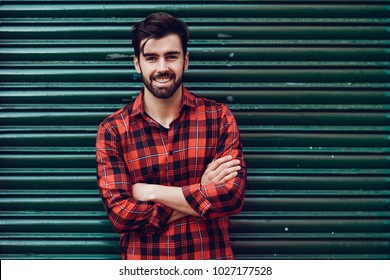 Young smiling man, model of fashion, wearing a plaid shirt with a green blind behind him. Guy with beard and modern hairstyle in urban background. - Powered by Shutterstock
