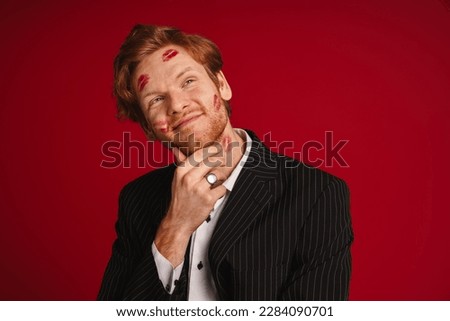 Young smiling man with red lipstick marks on his face touching his chin and looking away isolated over red studio wall