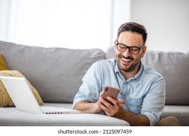 Young smiling man relaxing using a laptop is doing a video conference meeting at home. A young creative man looks at the screen and types a message with a smartphone. the concept of working from home