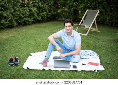 Young Smiling Man Sits On Picnic Blanket On The Backyard And Working On Laptop